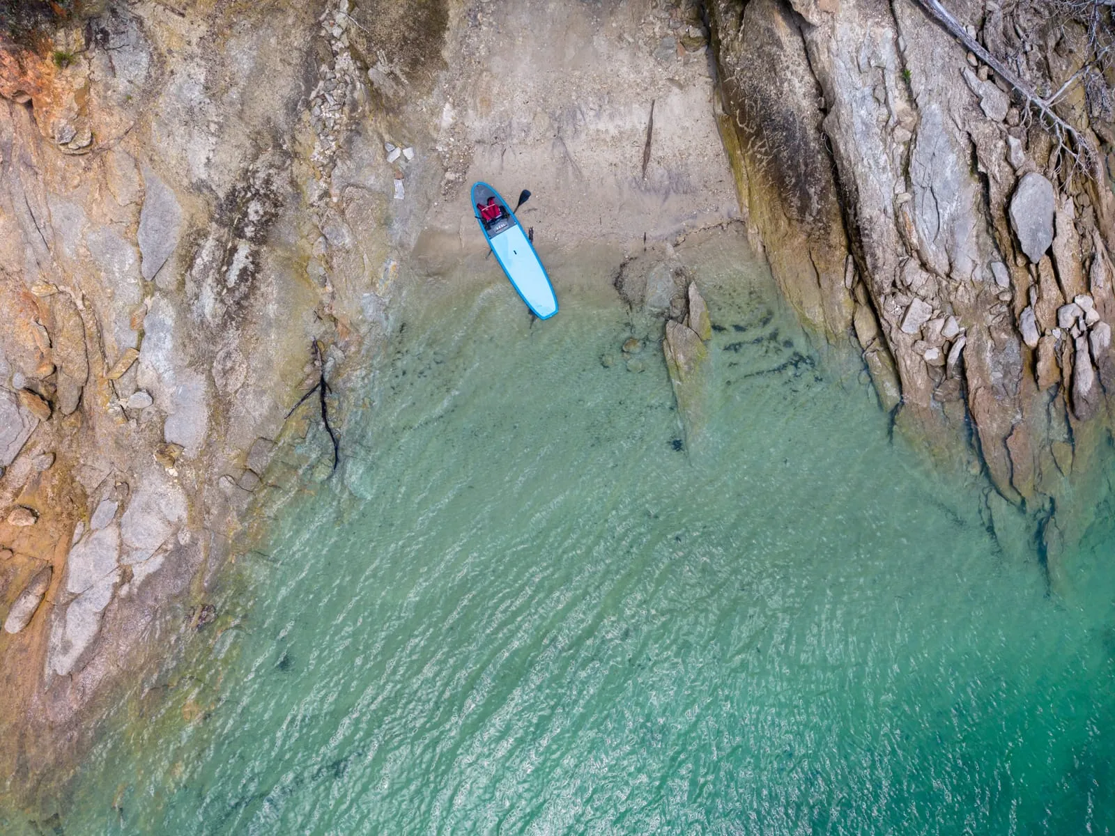 Overhead drone view of a blue paddleboard pulled onto a small sand beach at Lake Jocassee, with clear green water and rocky shoreline on both sides.