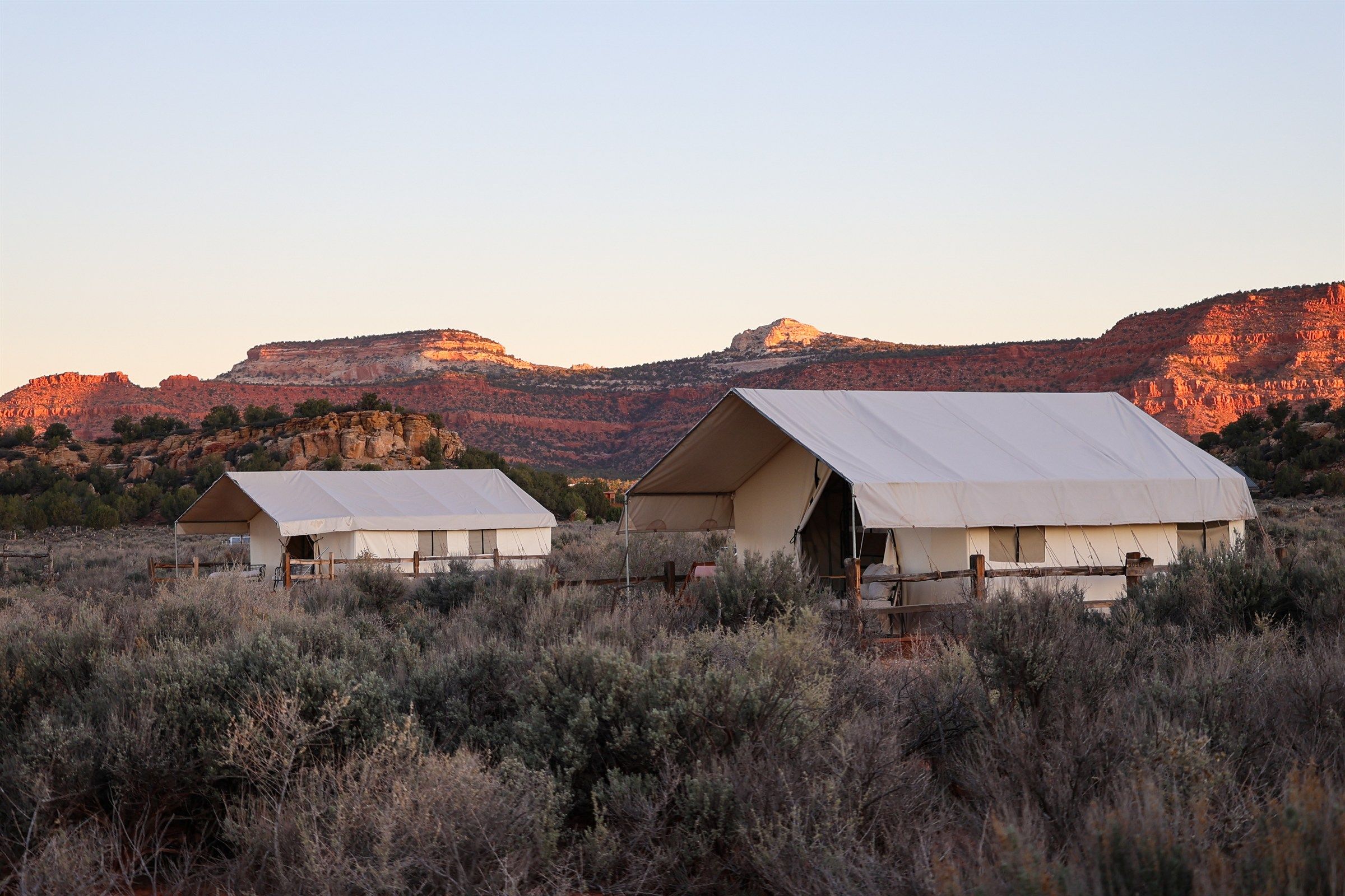 Two canvas glamping tents in sagebrush at sunset, with red sandstone cliffs lit gold behind them in southern Utah.